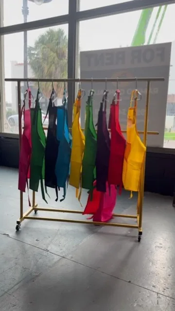 A rolling rack of brightly colored aprons in pink, green, blue, yellow, and red, waiting by the studio window.