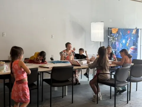 Children gathered around a long table, drawing and working with a mentor at the head.