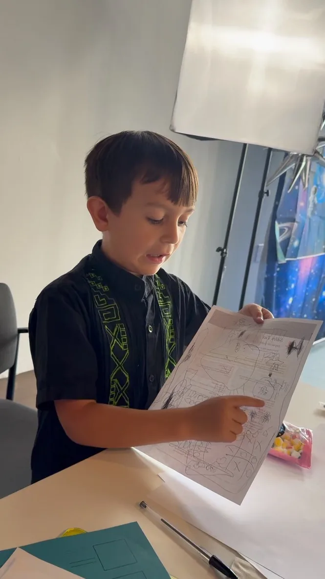 A boy holds up a page covered in his own drawings and ideas, talking through it.