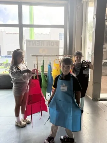 Three children pick out their aprons from the rack, one already wearing a blue one.